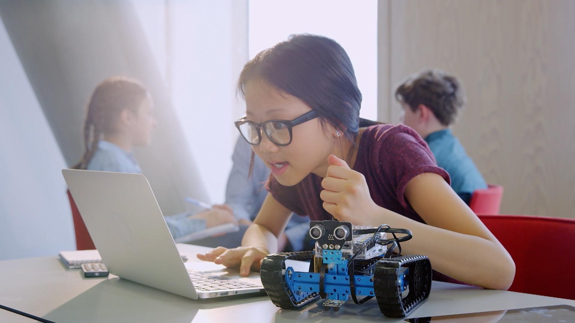 An Asian girl wearing glasses uses a white laptop. Next to her is a blue and black robot on wheels. 