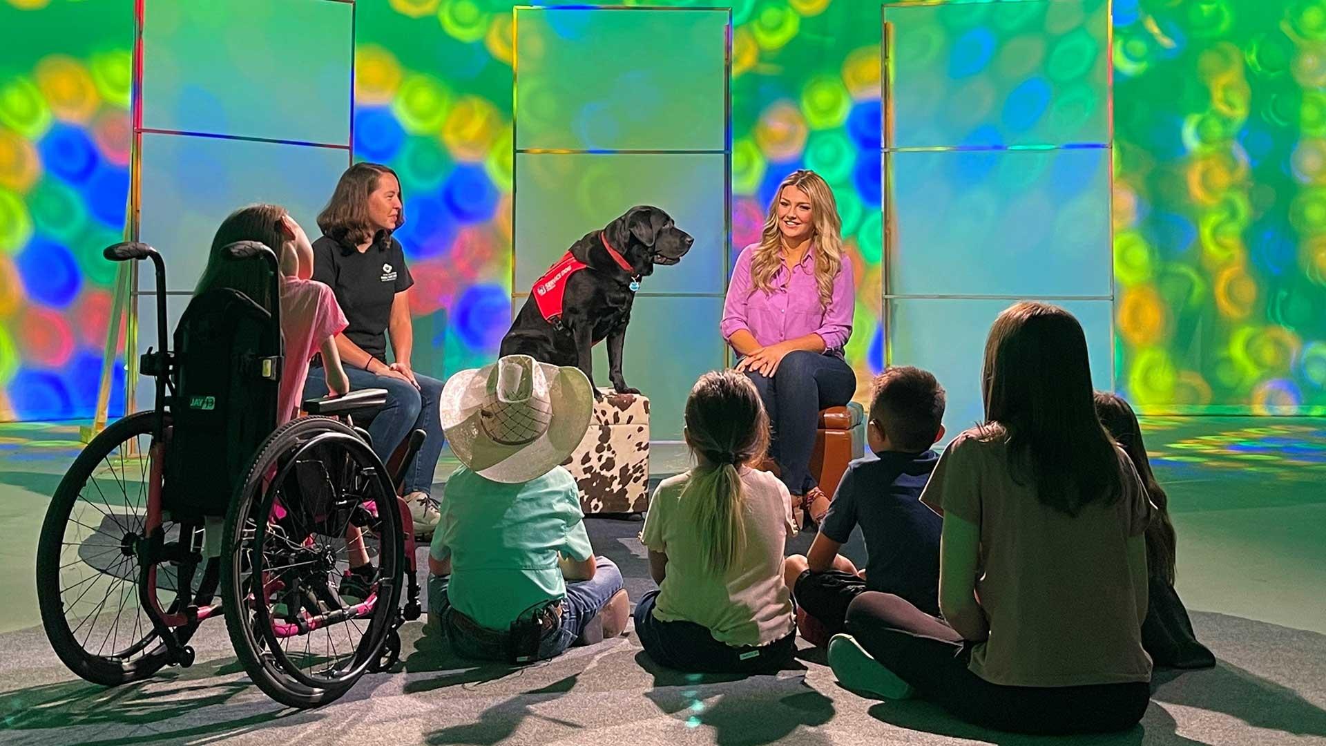 A blonde woman and a black service dog smile as they sit in front of a group of children. 