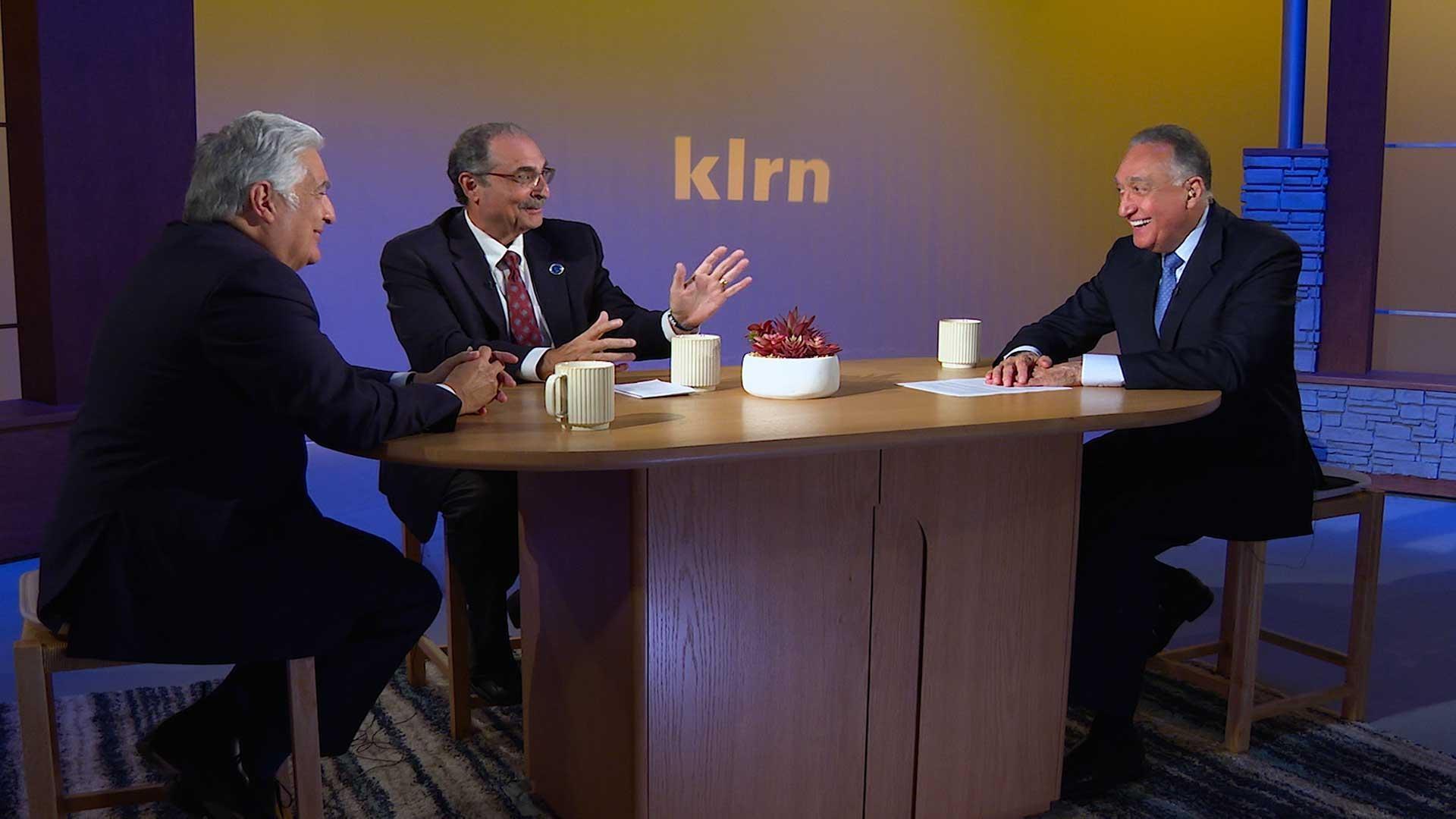 Three men sit at a desk, smiling and laughing. Behind them a blue-ish, purple background with the letters 'KLRN' appear
