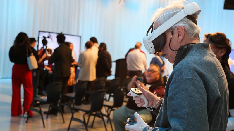 A person wears a VR headset while holding controls in their hands in a crowded room