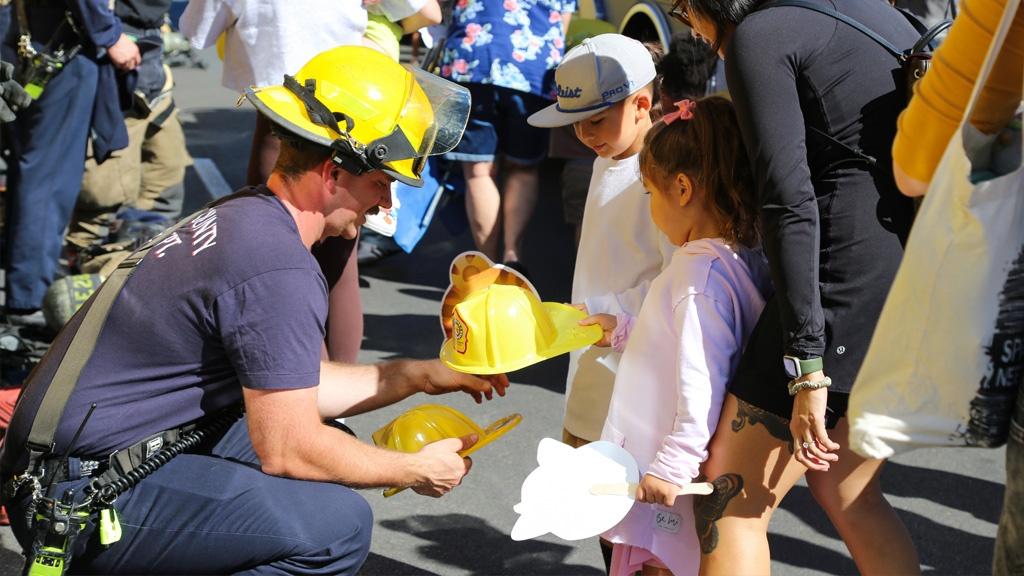 A firefighter crouches down as he hands a yellow helmet to a child