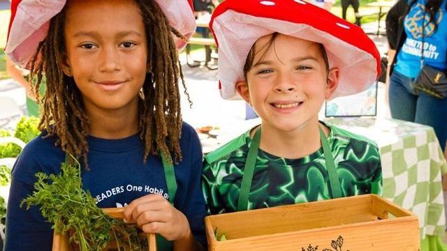 Two children wearing red and white mushroom hats and green aprons as they hold containers of freshly picked vegetables