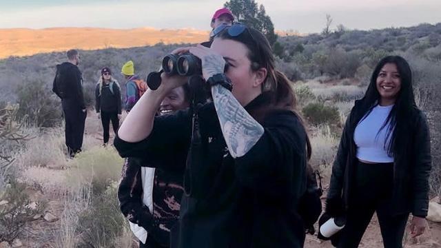 A person looking off to the left of the camera holding binoculars while a woman smiles as she looks on, both standing in a desert landscape with early morning sunrise light illuminating the hills behind them