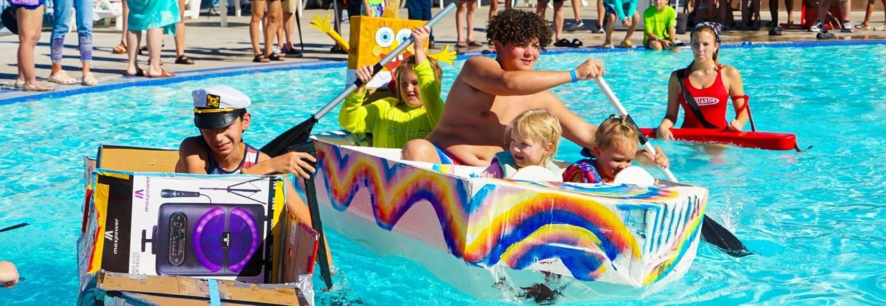 Children paddle cardboard boats afloat in the blue water of a public pool as a lifeguard in a red one-piece swimsuit looks on