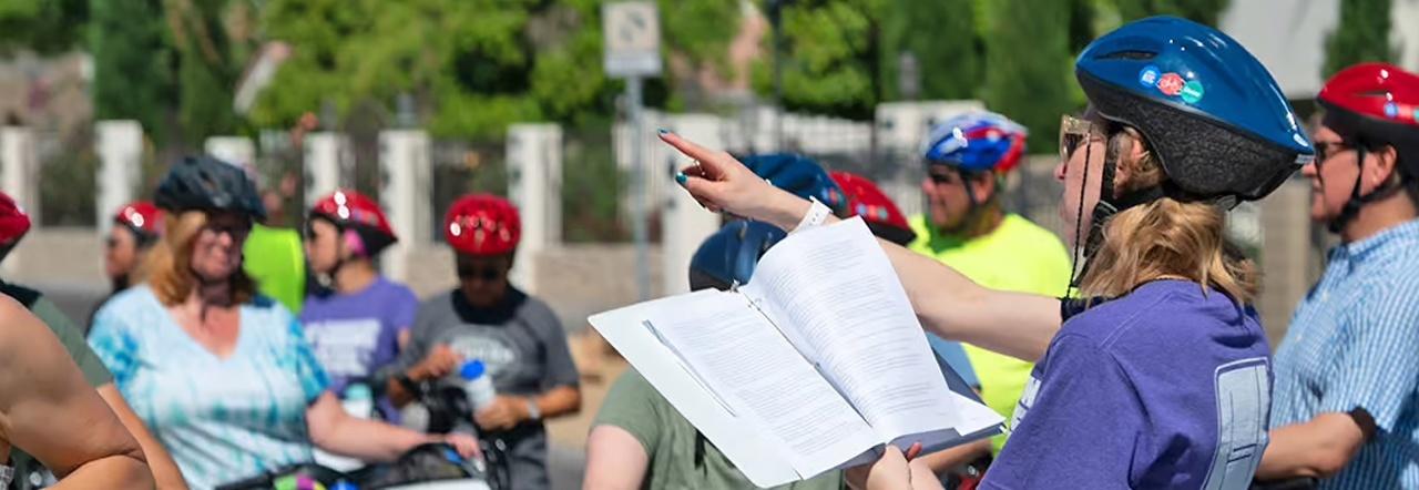 A tour guide wearing a purple t-shirt and a blue bike helmet looks at a three-ring binder of typed notes as they point in the distance in front of a crowd of bicyclists