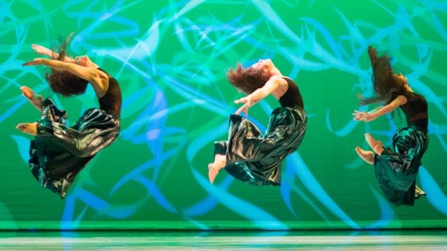 Three dancers arching their bodies as they leap through the air against a green backdrop with blue swirling lights