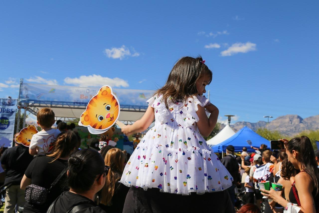 A child wearing a dress is perched on an adult's shoulders, carrying a fan with Daniel Tiger's face on it