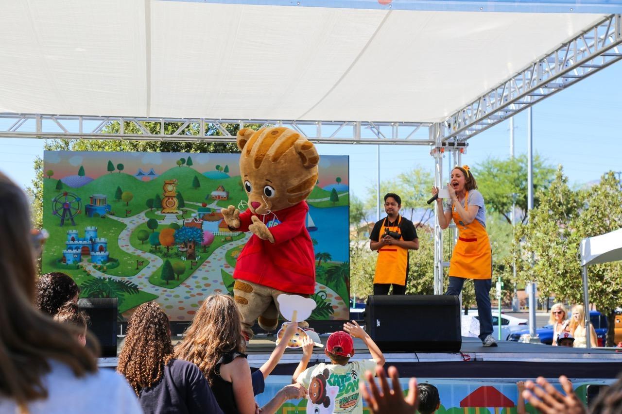 A Daniel Tiger mascot waves to the audience from an outdoor stage as two adults in PBS KIDS aprons speak into microphones