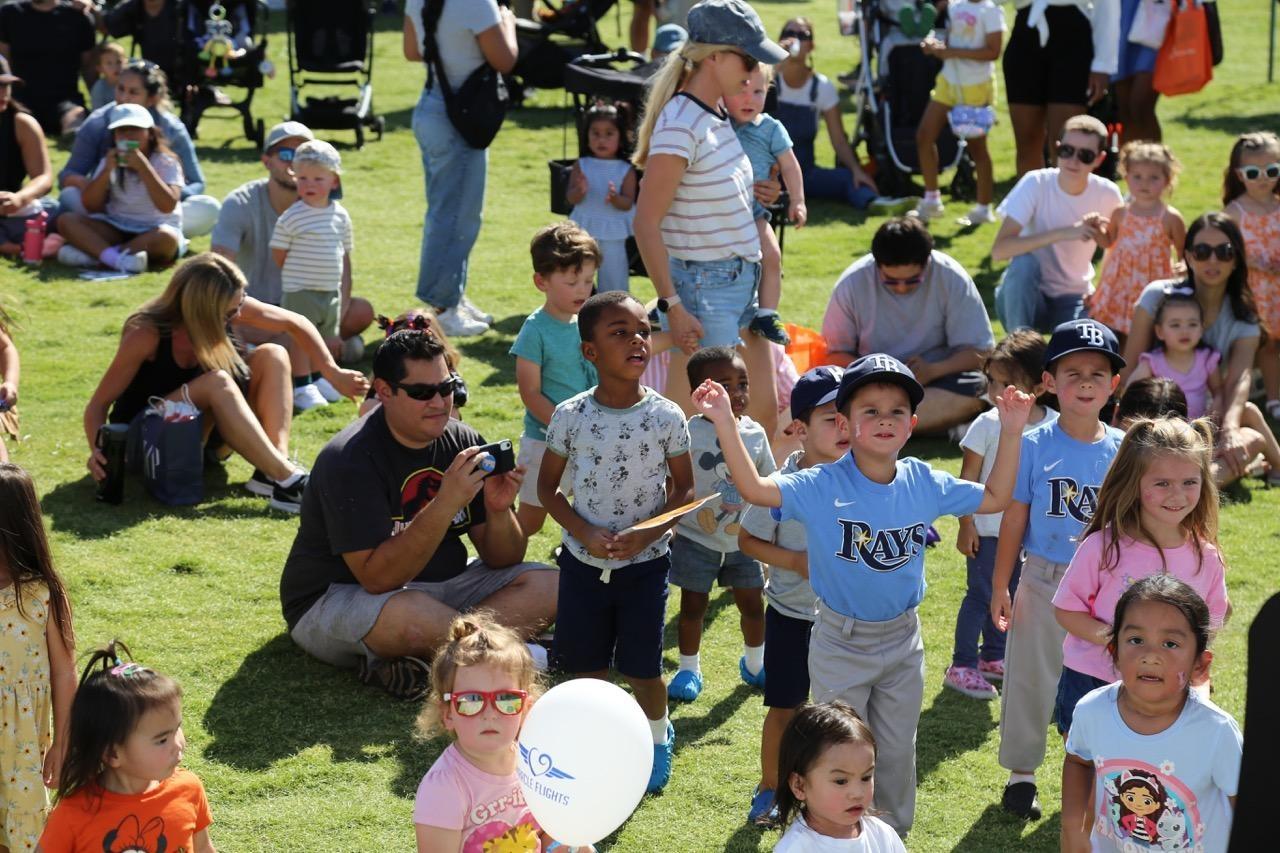 Children stand at attention with adults sitting on a grassy lawn outdoors