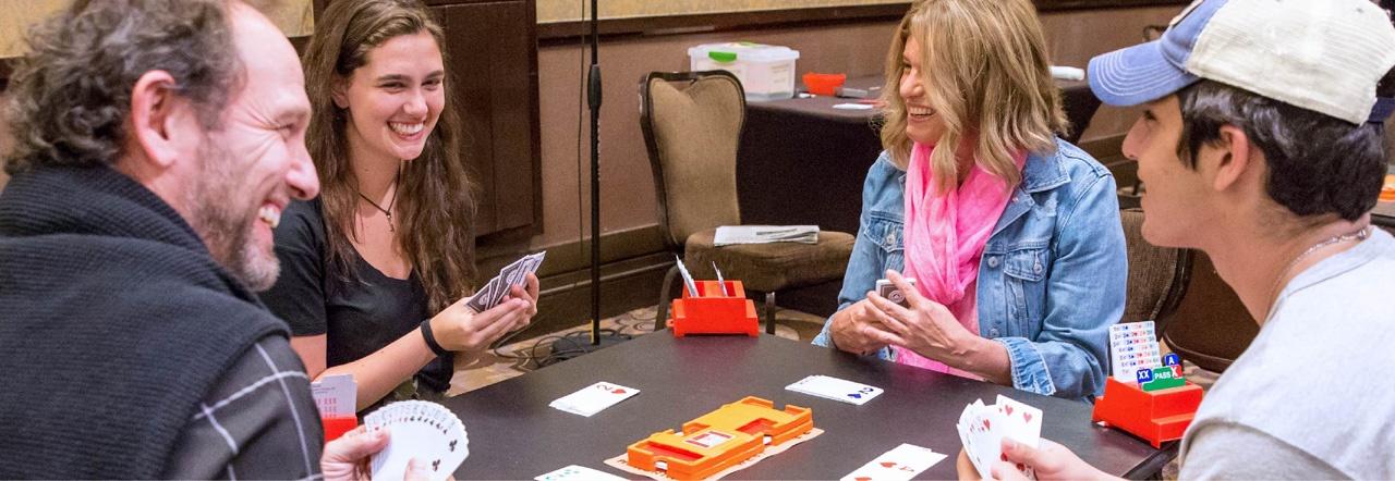 Four people sit around a square table smiling as they hold playing cards in their hands