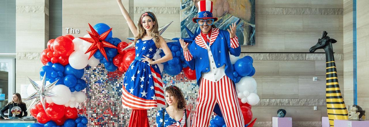 Performers dressed in patriotic garb pose in front of red, white, and blue balloons inside City Hall