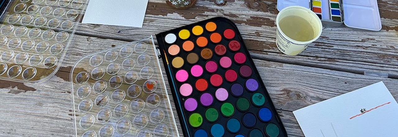 Watercolor paints and a cup of water spread out on a wooden table