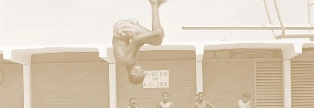 A sepia tone photo of an African American child jumping off a diving board into a public pool while other children watch