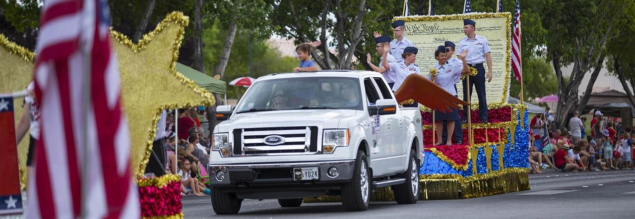 Military personnel ride a parade float pulled by a white truck down a paved street lined with spectators