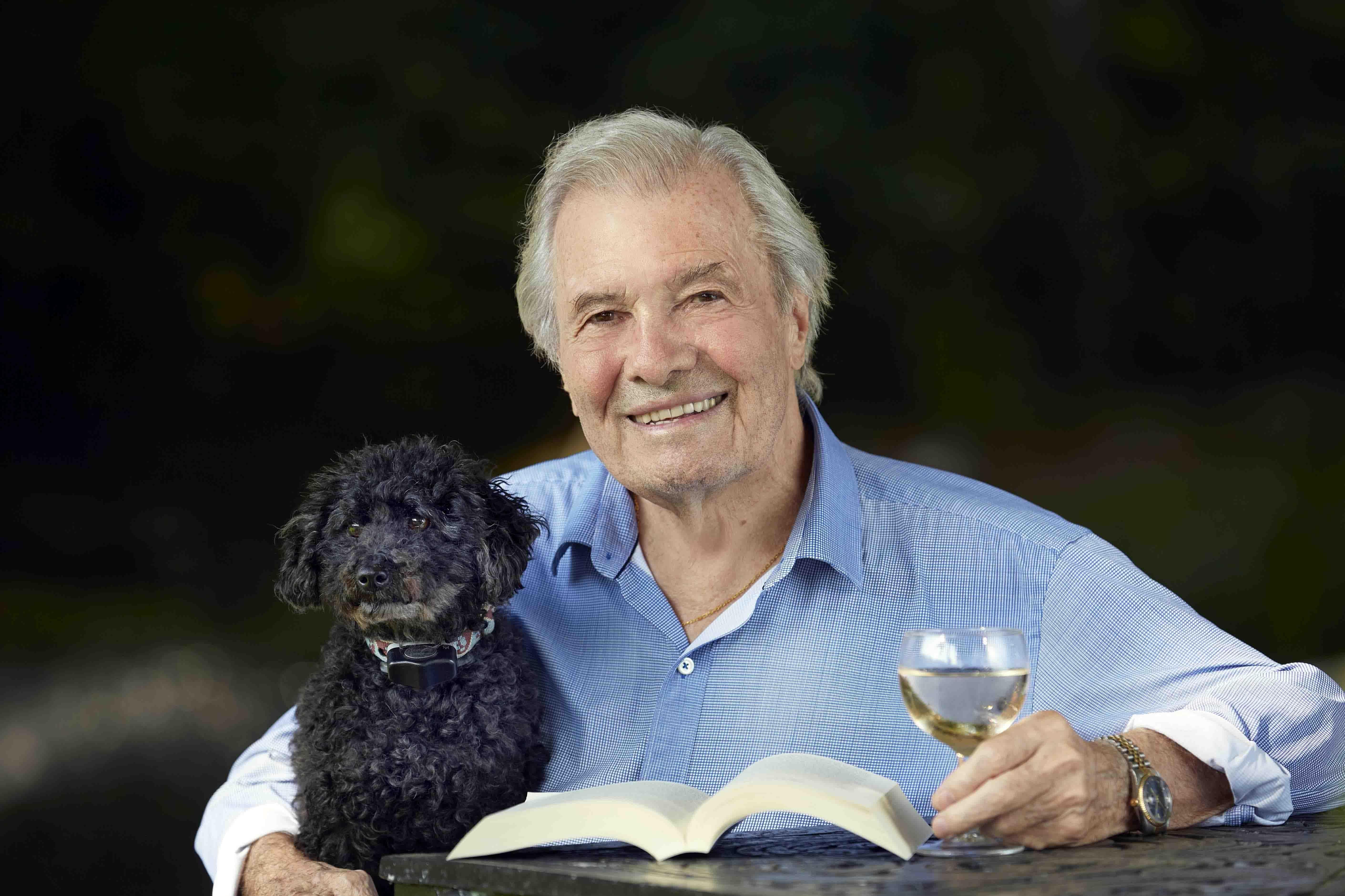 Jacques Pepin holding a glass of white white in his left hand while sitting at a table with an open book in front of him and his right arm around a curly-haired black dog