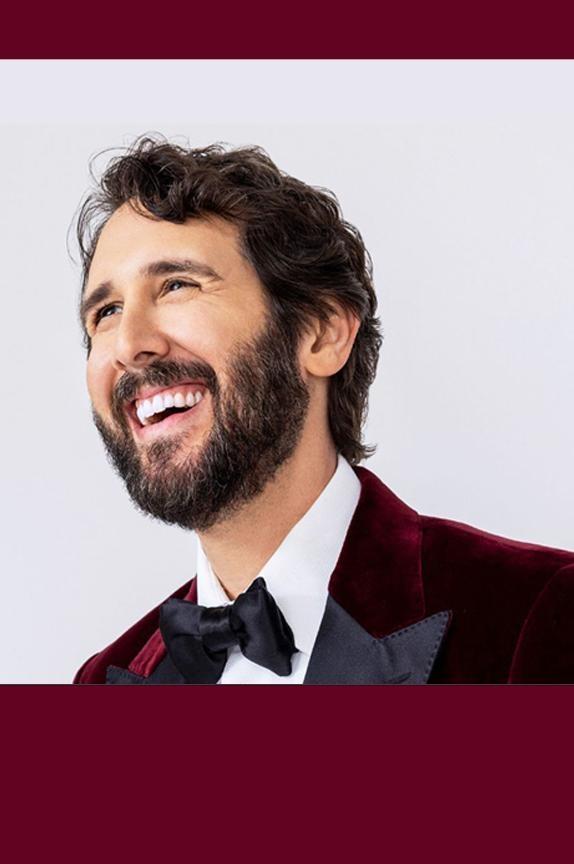 Josh Groban smiling off to the side while wearing a burgundy velvet tuxedo with black lapels