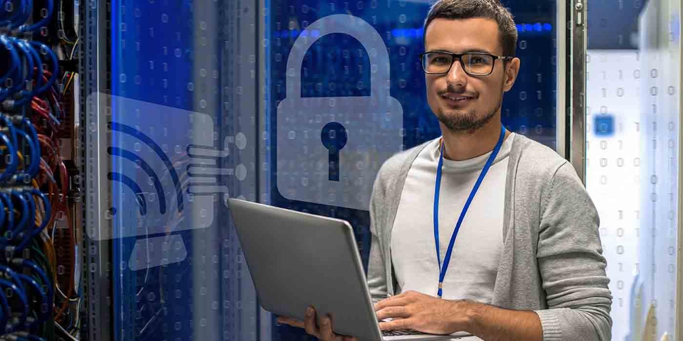 A person wearing glasses, with short dark hair and a dark goatee, looks at the camera while holding a silver laptop in their right arm and typing with their left, set against a backdrop showing binary codes and graphics of computer networks and a padlock