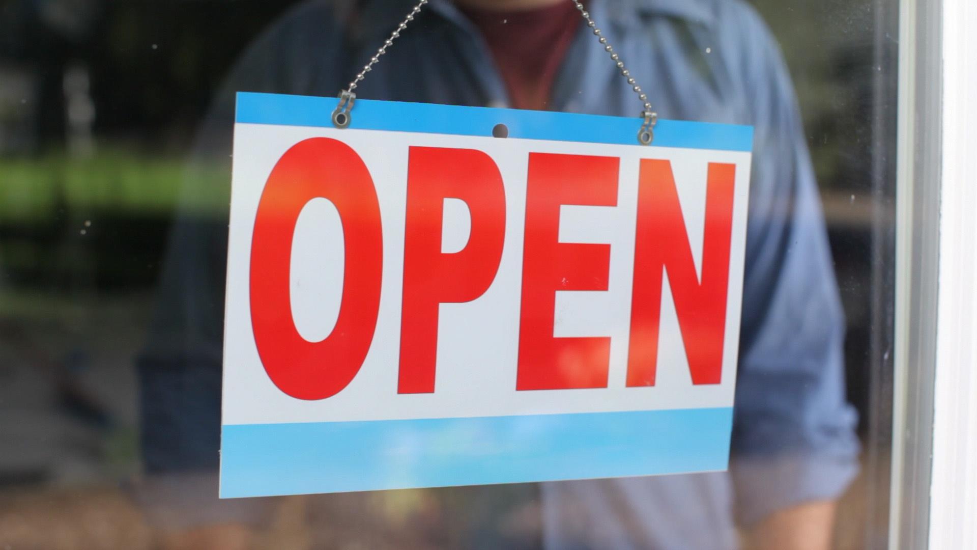 "OPEN" sign with red letters hanging in a window with a reflection of a person wearing a light blue button-down shirt