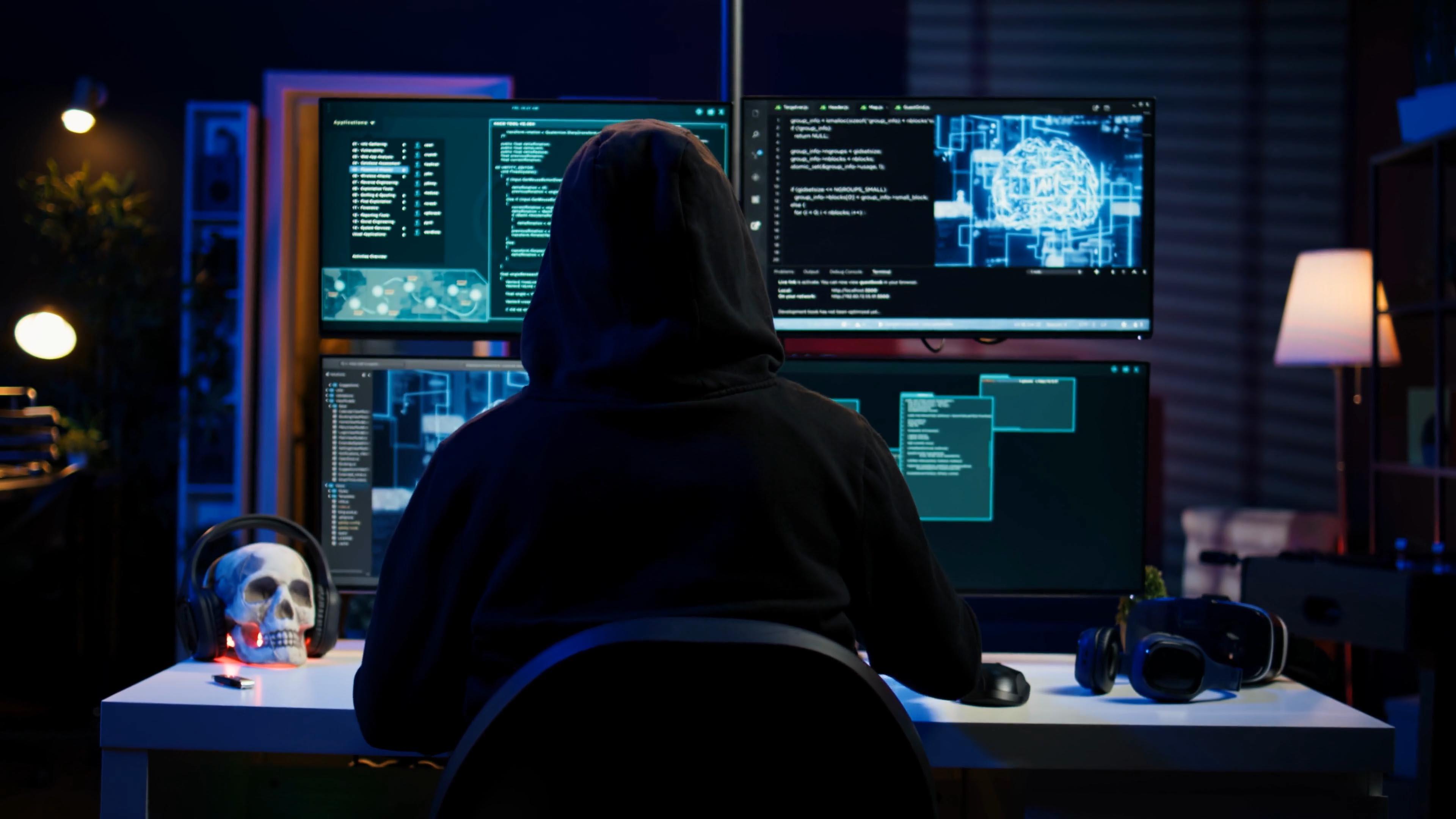 A hacker wearing a hoodie sits at a desk facing several computer screens in a dark room