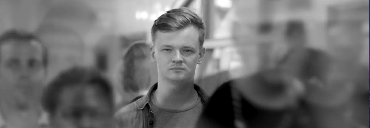 Black and white photo of a light-haired young man standing in a crowd of blurry figures, looking straight at the camera