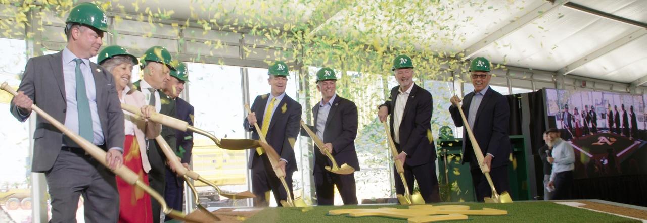 A line of seven men wearing suits and one woman wearing a red skirt hold golden shovels over a dirt platform while wearing green hard hats emblazoned with the logo for the A's baseball team