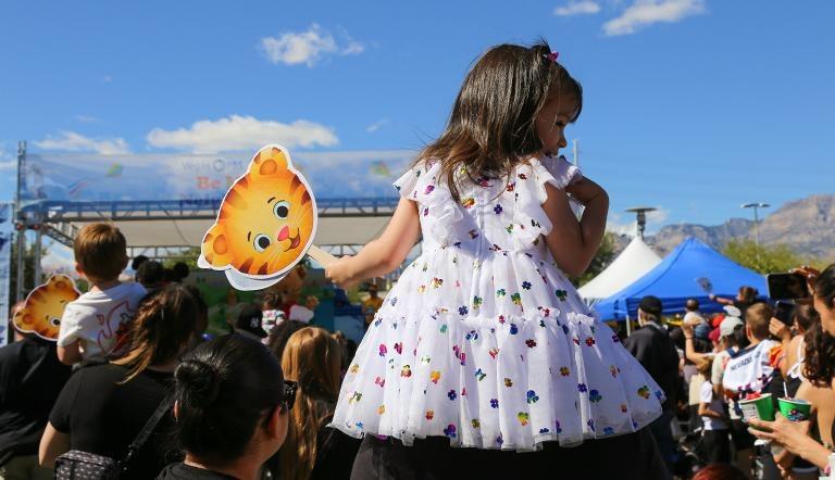 A child wearing a fluffy dress looks out over an outdoor festival while holding a Daniel Tiger fan
