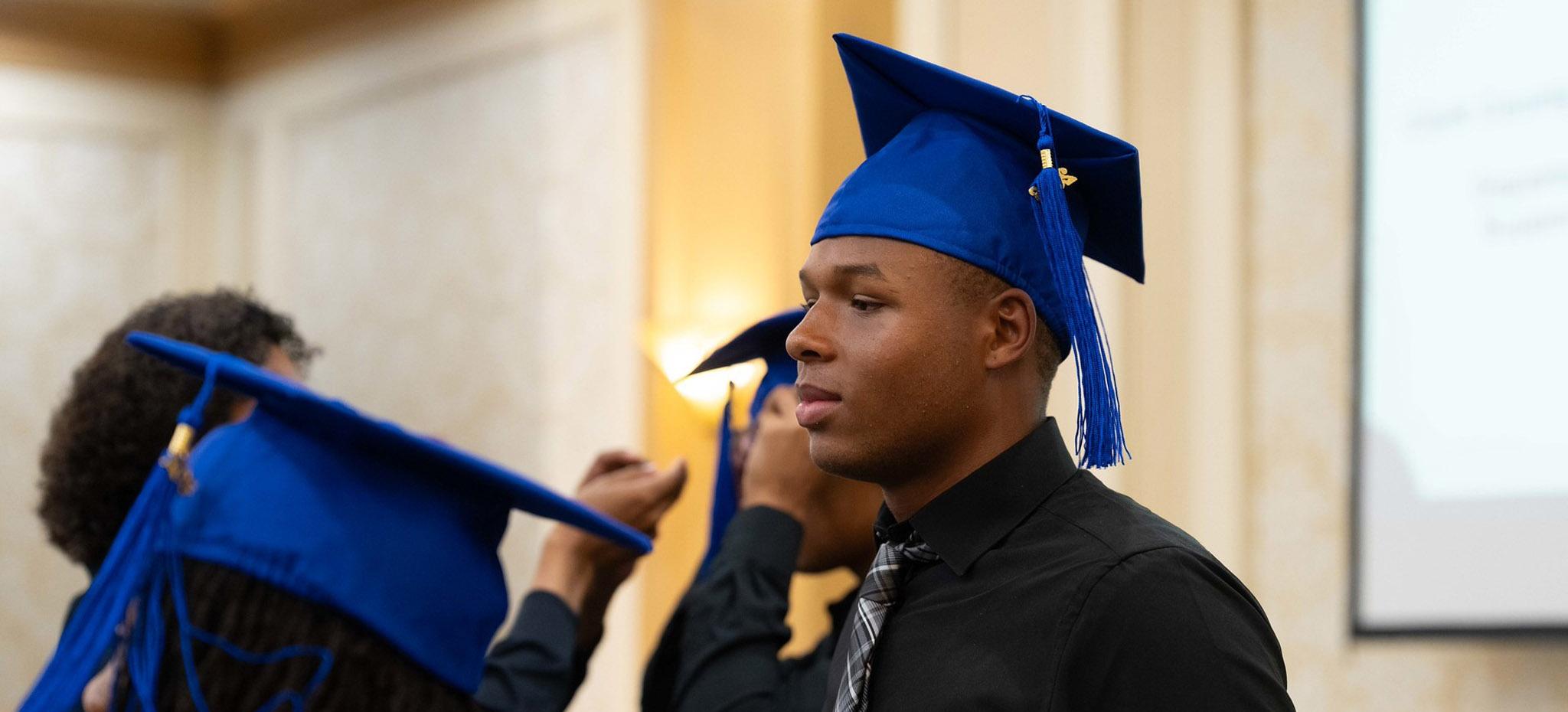 A graduate in a black dress shirt and striped tie looks down while wearing their blue graduation cap and tassel