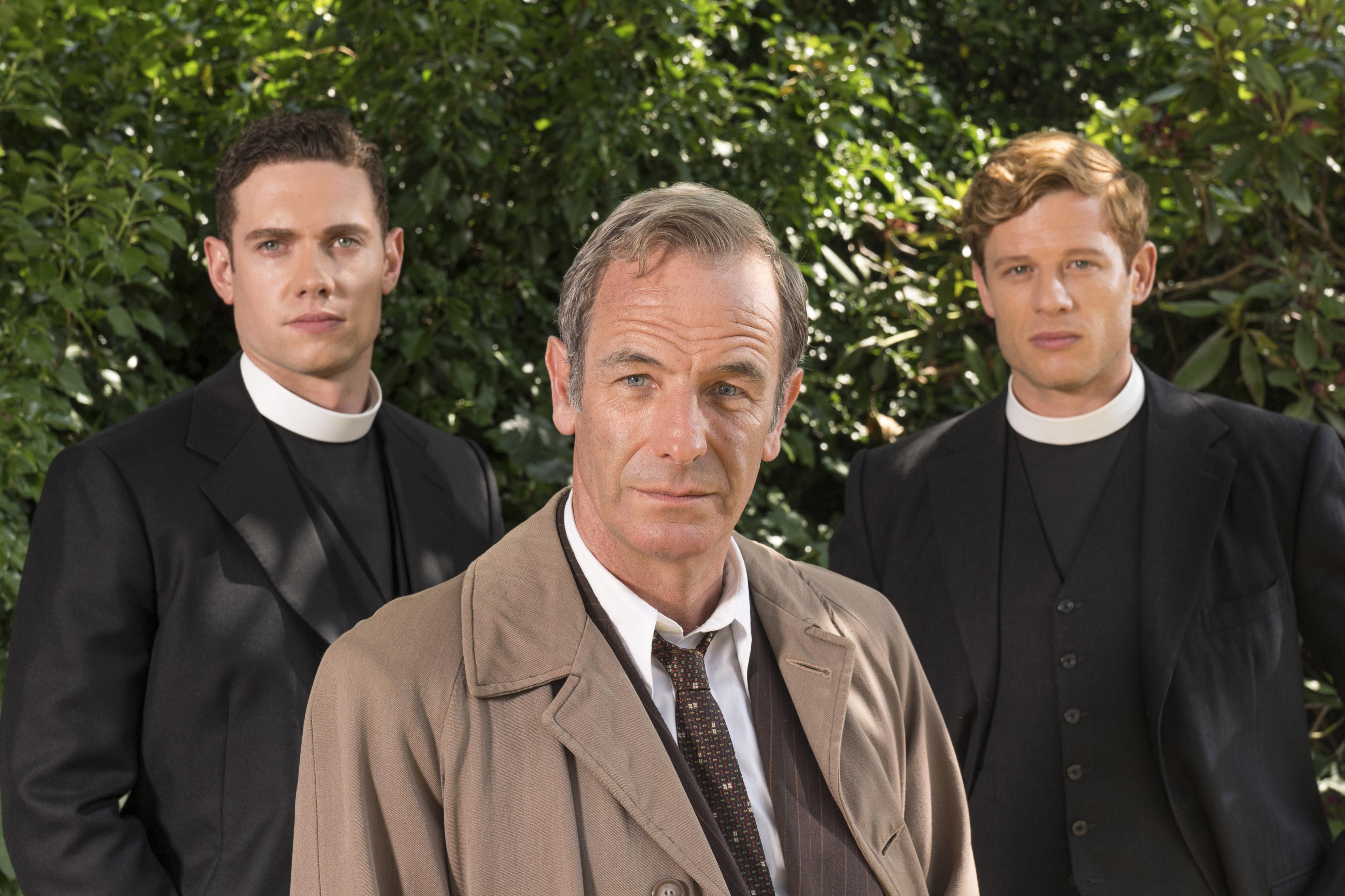Three men, including two clergy members, stand outdoors in front of greenery.