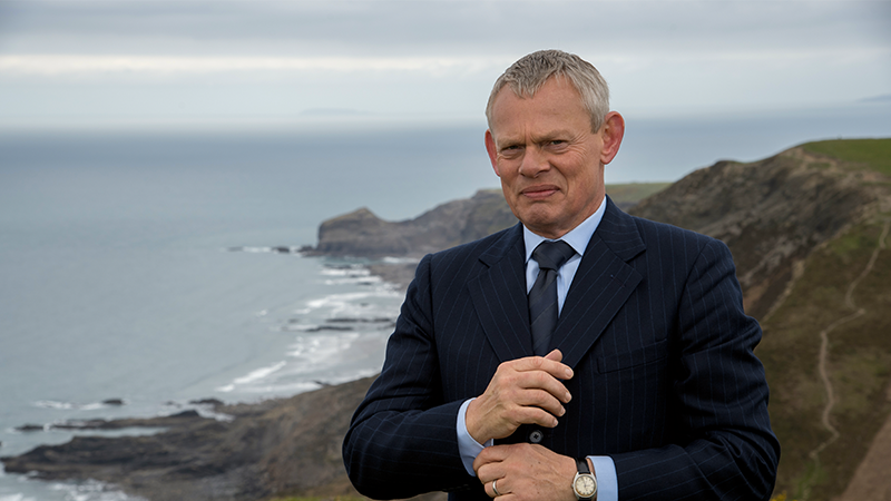 Doc Martin standing in a suit adjusting his cuffs in front of a cliffside