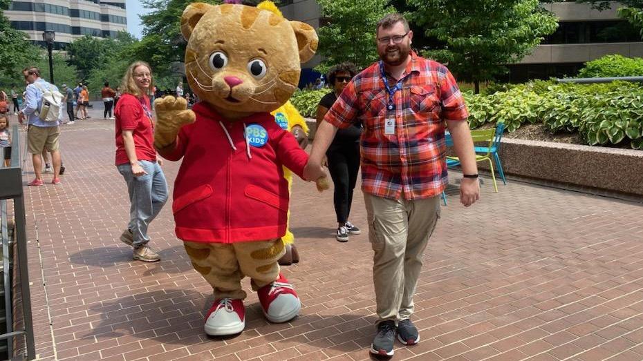 A mascot of Daniel Tiger is holding hands and walking with a student employee.