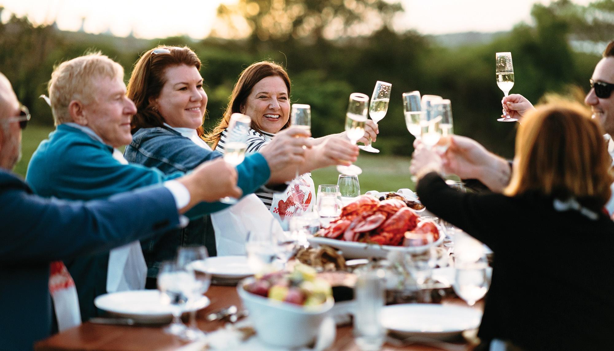 A group of people sit around a table with lobster, holding up their drinks to one another.