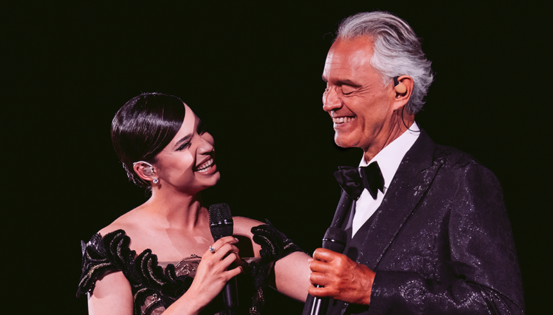 Andrea Bocelli and Sofia Carson holding microphones, smiling at each other.