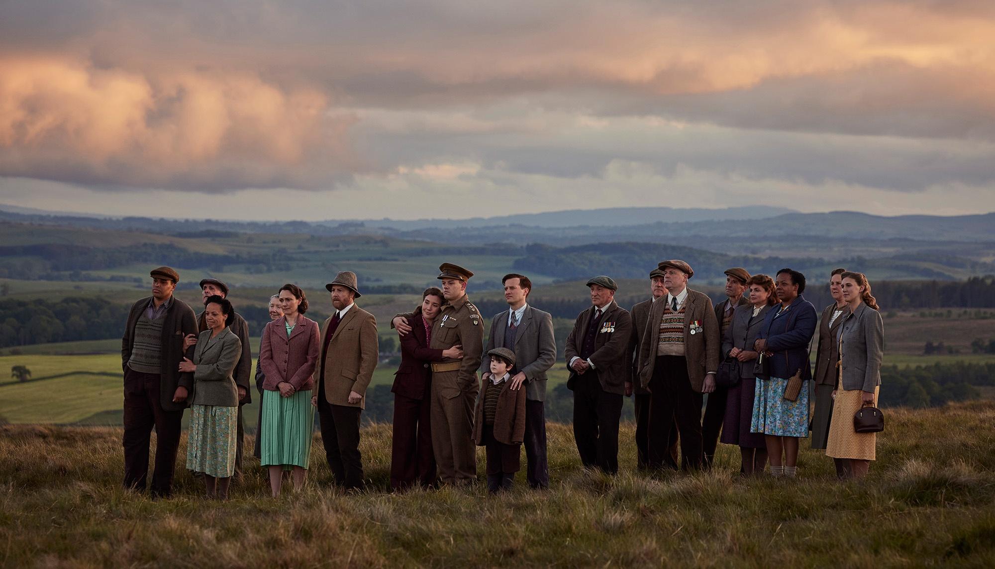 The cast of All Creatures Great and Small stand together on top of a field.