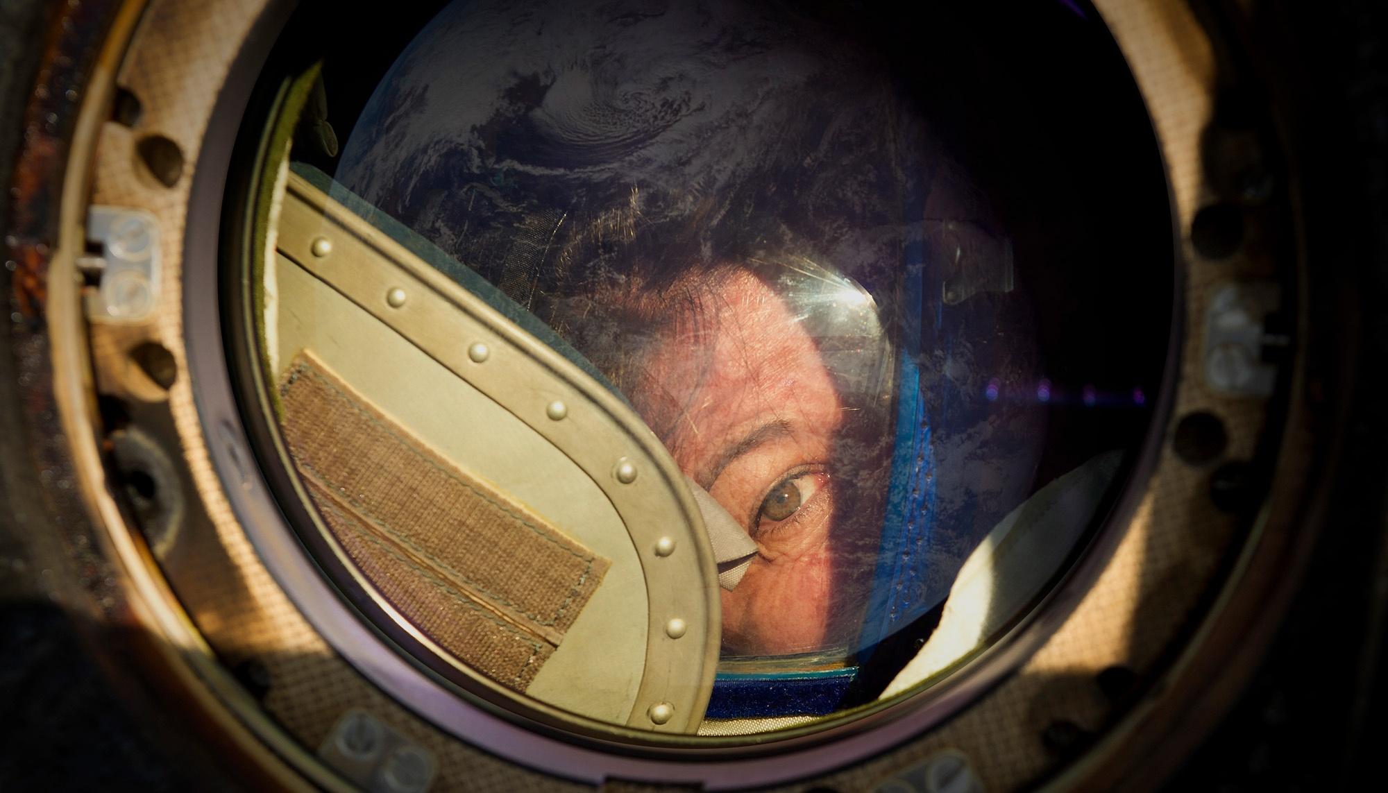 An astronaut looking out of a window in space with Earth reflecting off of the glass.