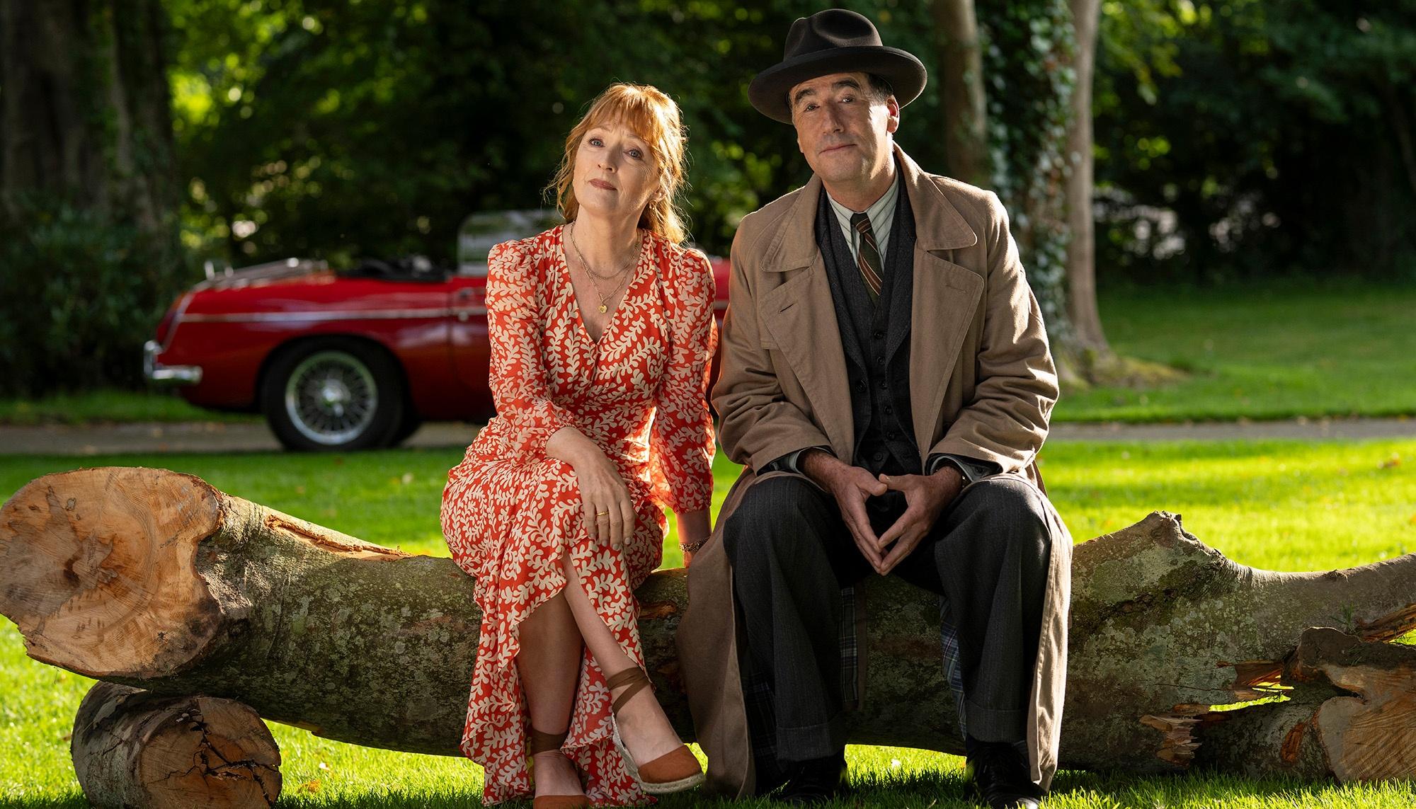 Characters Susan Ryeland and Atticus Pund are sitting together on a log.