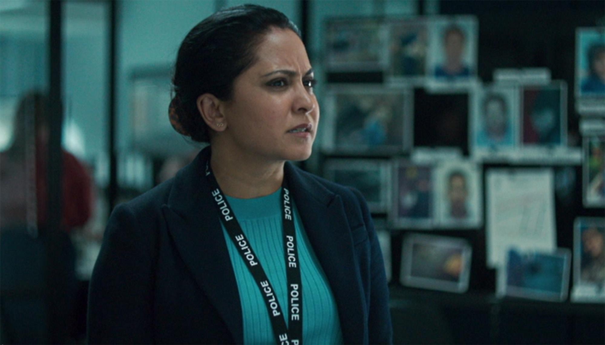 Character Rachita Ray is standing in front of a bulletin board with suspect pictures, wearing her police lanyard.