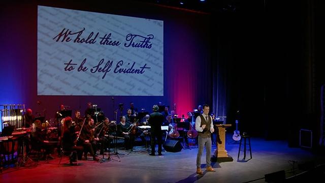 A live musical concert stage with many different musicians on their instruments and a man standing in front telling a story