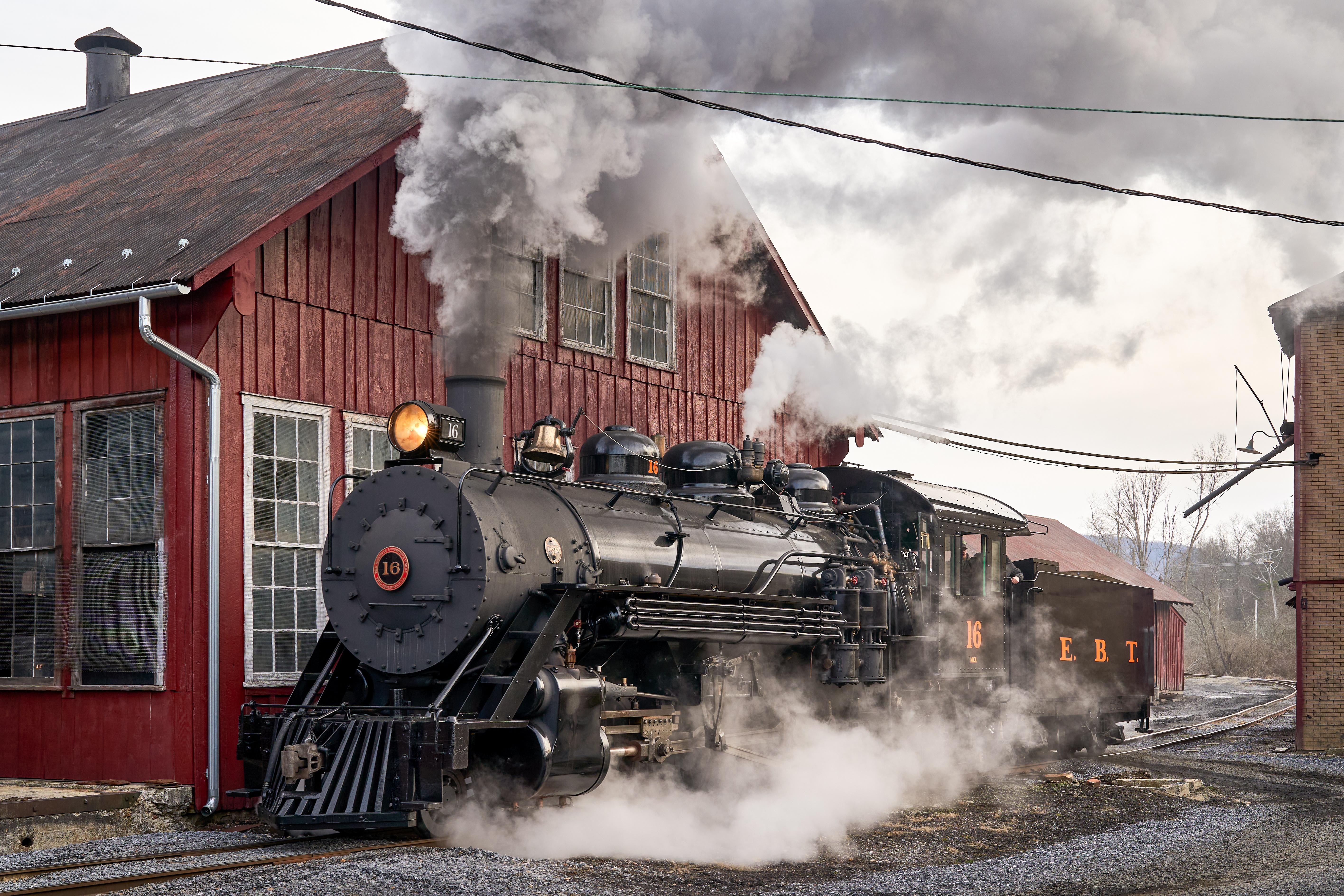 Black steam locomotive stands beside a large red wooden rail building, billowing thick white steam and smoke into the air as it appears to be preparing to depart or has just arrived at a historic train yard.