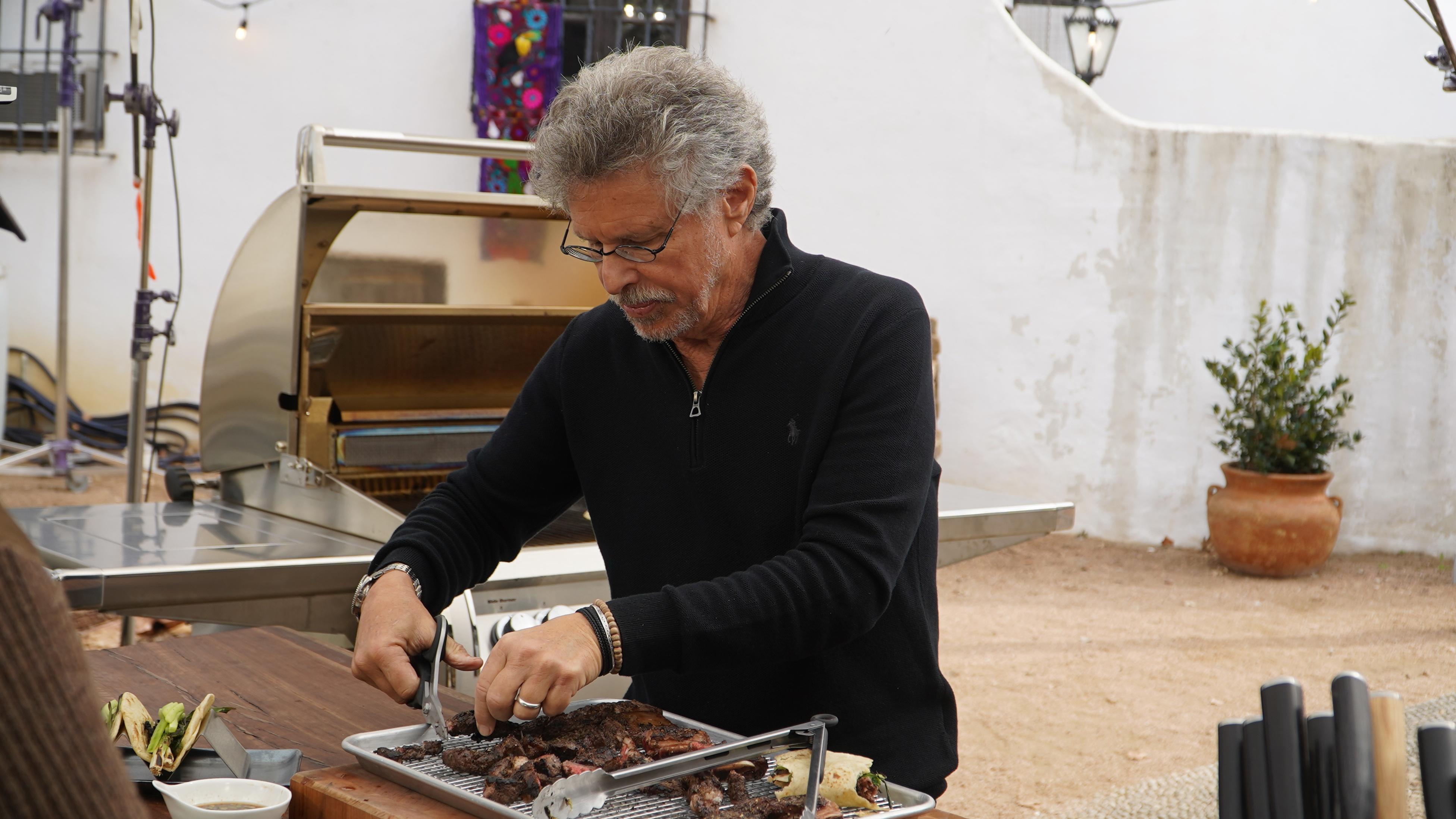 Steven Raichlen shredding Barbecue with a kitchen scissors by a grill outdoors.