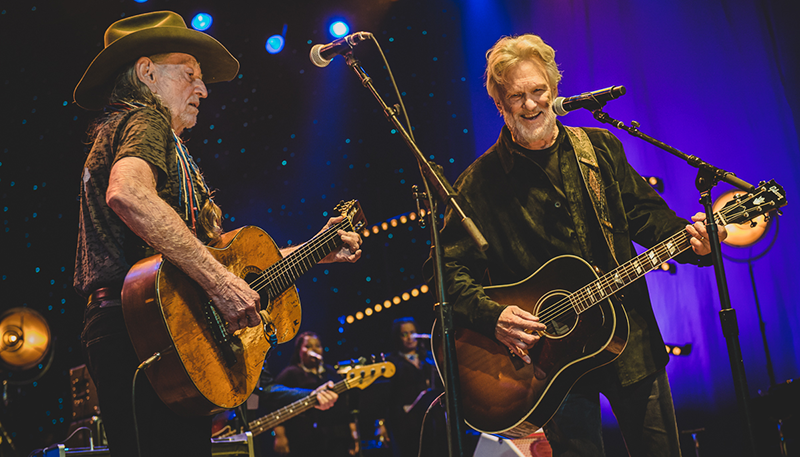 Kris Kristofferson playing the guitar on stage while singing into the microphone with another musician.
