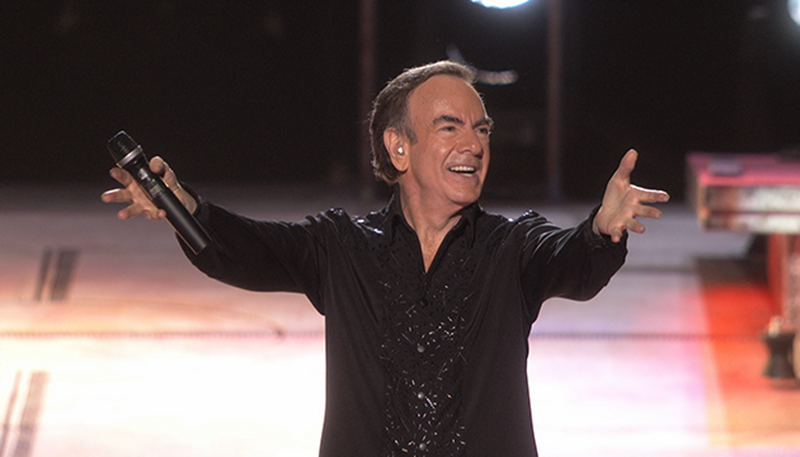 Neil Diamond on stage with his hands spread out holding a microphone in one hand, smiling.