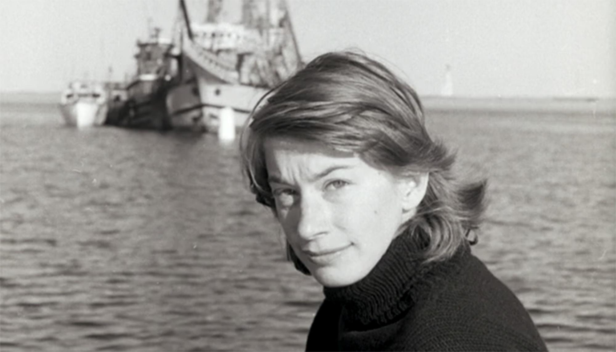 A black and white image of Mary Oliver looking at the camera while behind her lies the ocean