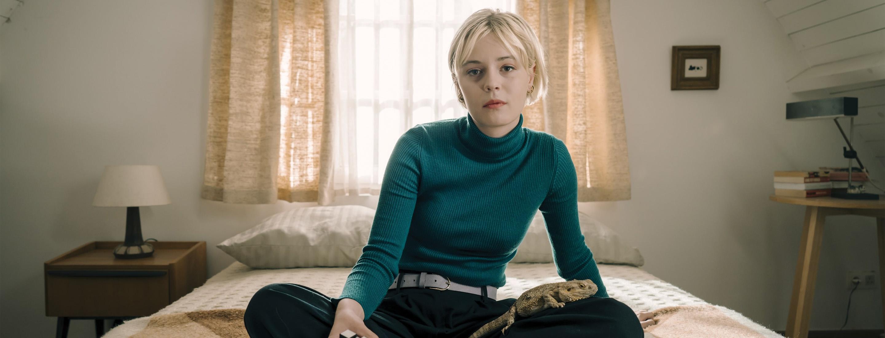 Patience sits on a bed with a lizard in a softly lit bedroom.