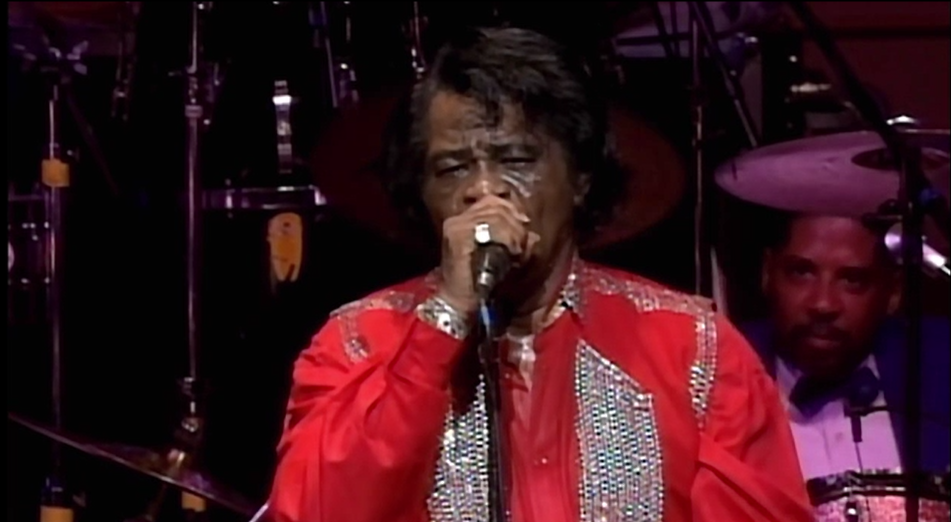 James Brown dressed stylishly in a red suit holding a microphone at a live band performance.