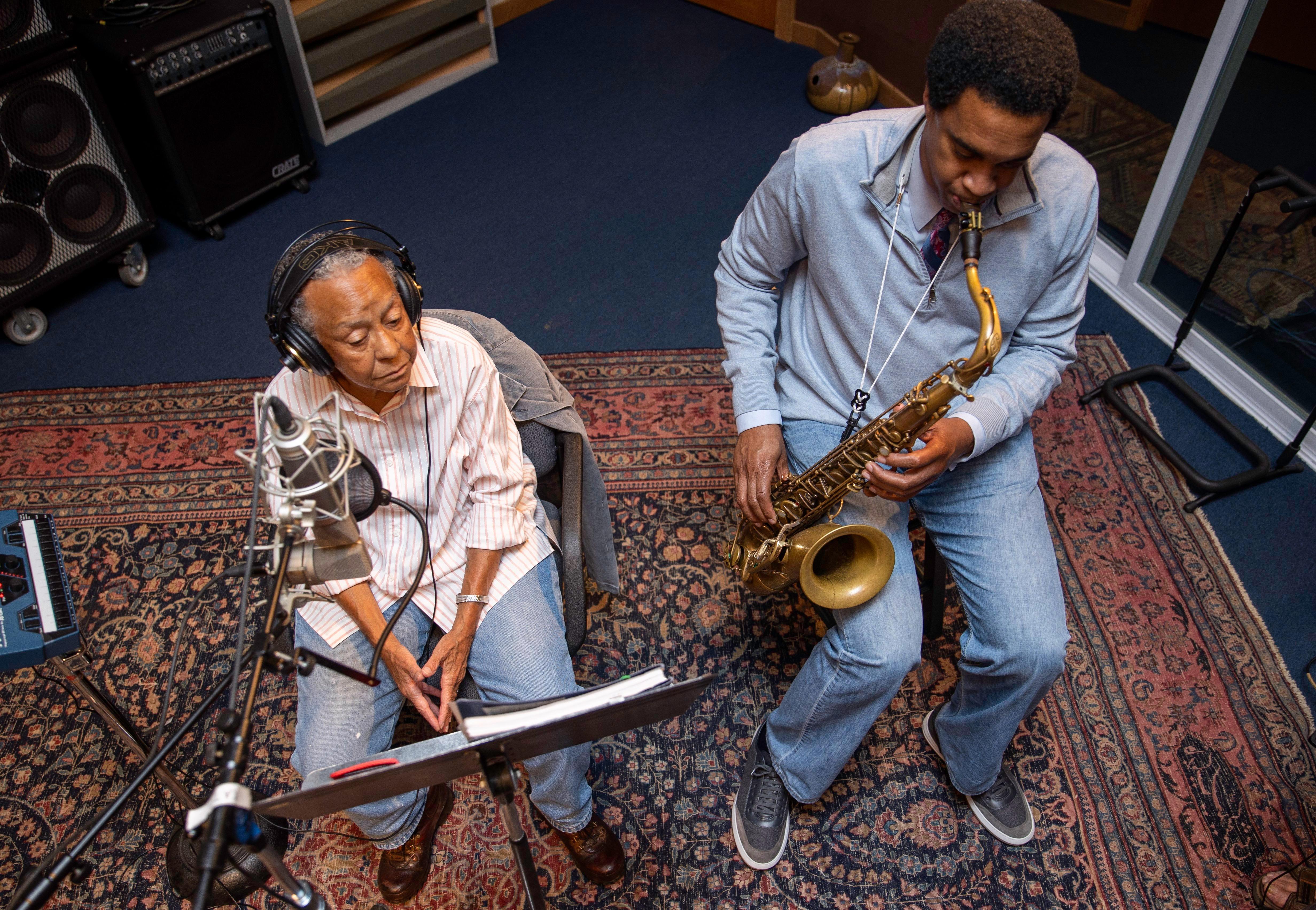An overhead shot shows poet Nikki Giovanni sitting next to Javon Jackson in a recording studio as he plays the saxophone.