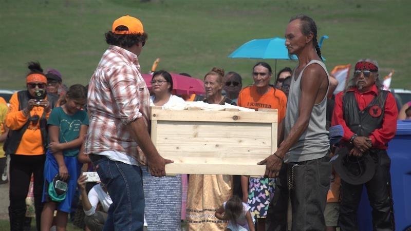 Two men carry a small pine casket, there are many people standing in the background watching the procession.  