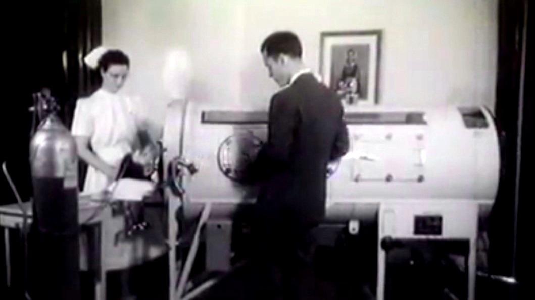 Archival photo of a nurse and doctor standing next to an iron lung with a child in it. 