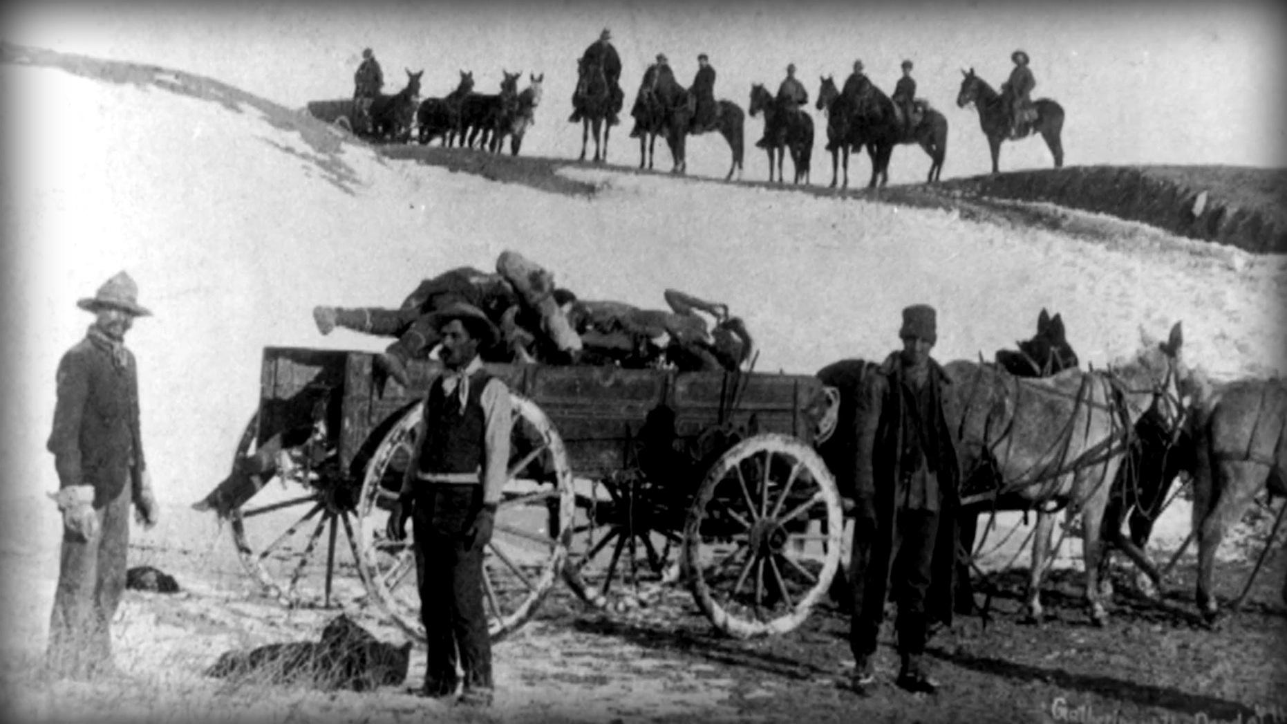 Archival image of three soldiers at the Wounded Knee Massacre site loading Native American bodies onto a horse drawn wagon.  Additional soldiers are on horseback in the background. 