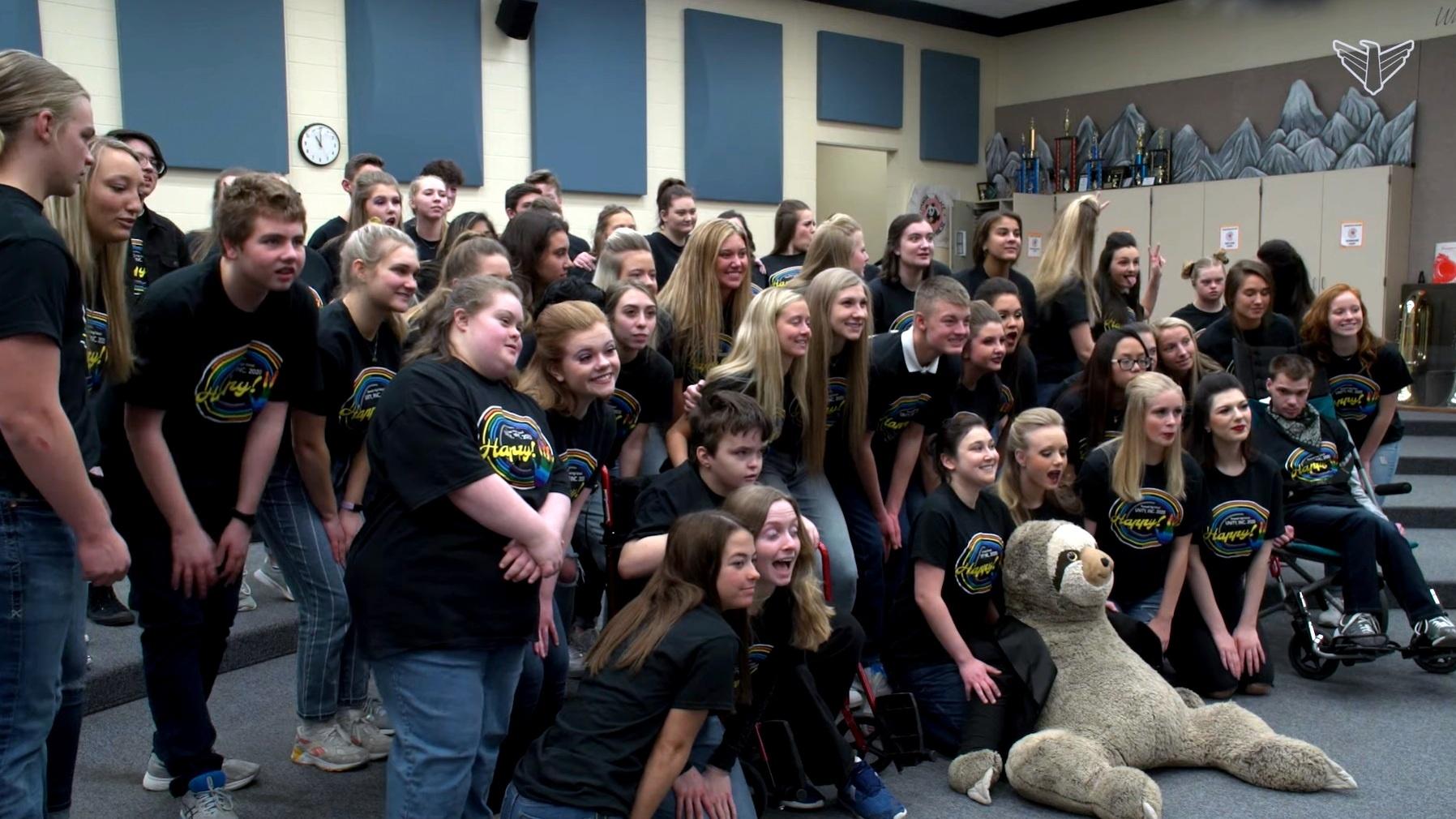 A group of students is taking a photo in a music classroom with a giant stuffed sloth in front of them on the floor. 