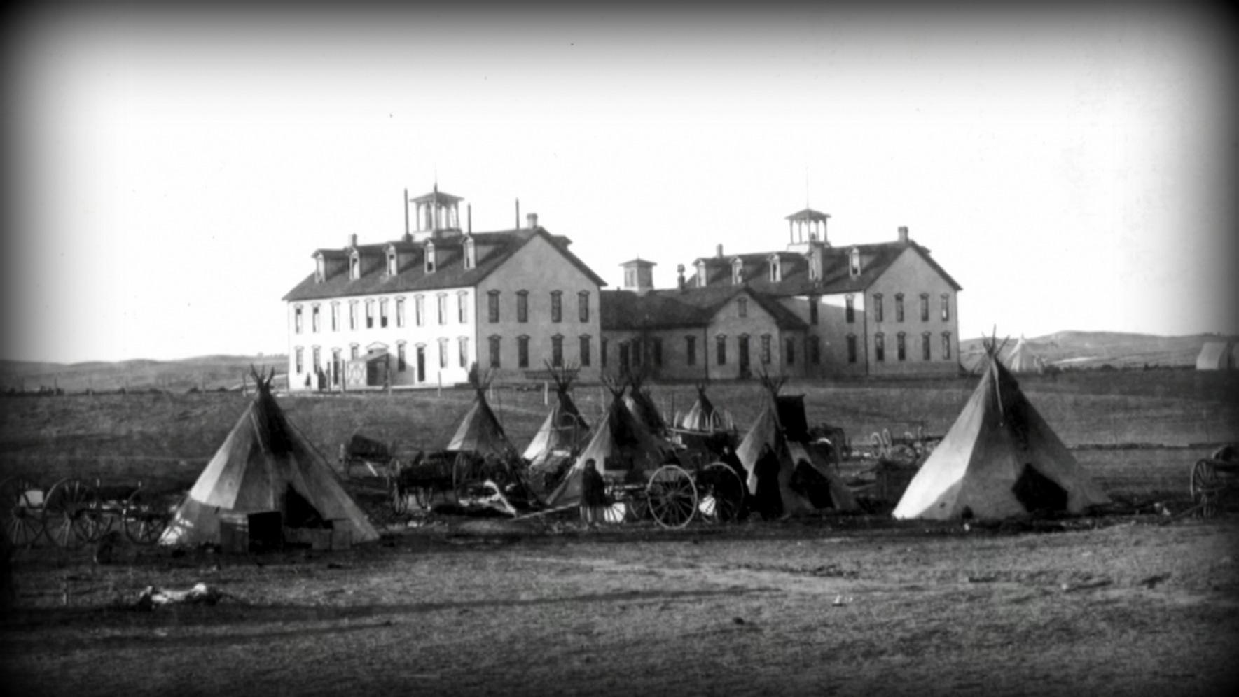 Archival photo of eight teepees set up in a camp in front of three buildings in the background. 